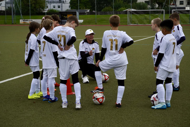 Ein Fußballtrainer spricht auf einem Sportplatz im Halbkreis zu einer Gruppe von Kindern, die in weißen Trikots mit der Aufschrift „Herbstcamp 2024“ gekleidet sind. Die Kinder hören aufmerksam zu, einige lehnen sich auf ihre Bälle. Die Szene zeigt eine Trainingsbesprechung bei bedecktem Wetter auf einem Kunstrasenfeld. Im Hintergrund sind Tore und eine Laufbahn zu sehen.
