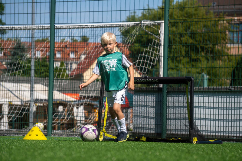 Ein blondes Kind im Vorschulalter spielt Fußball auf einem Kunstrasenplatz. Es trägt ein grünes Leibchen mit der Aufschrift „Unsere Amateure. Echte Profis.“ sowie weiße Sportkleidung. Vor ihm steht ein kleiner Fußball, daneben ein gelbes Hütchen und ein Mini-Tor. Im Hintergrund sind ein hoher Zaun, ein großes Tor und Gebäude zu sehen.