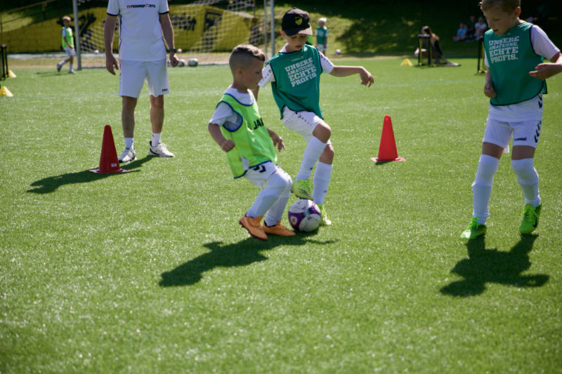 Mehrere Kinder spielen Fußball auf einem sonnigen Kunstrasenplatz. Zwei Jungen kämpfen im Vordergrund um den Ball, einer trägt ein grünes, der andere ein gelbes Leibchen. Im Hintergrund beobachtet ein Trainer in weißer Sportkleidung das Geschehen. Weitere Kinder, Hütchen und Tore sind auf dem Platz verteilt. Die Leibchen tragen den Aufdruck „Unsere Amateure. Echte Profis.“.