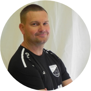 **Alt-Text:** Ein Mann in schwarzem Sportshirt mit dem Logo „Post SV Dresden“ steht seitlich vor einem hellen Hintergrund und schaut lächelnd in die Kamera. Auf dem Shirt ist auch sein Name mit Initialen „D.H.“ zu sehen. Die Pose mit verschränkten Armen wirkt freundlich und selbstbewusst – typisch für einen Fußballtrainer.