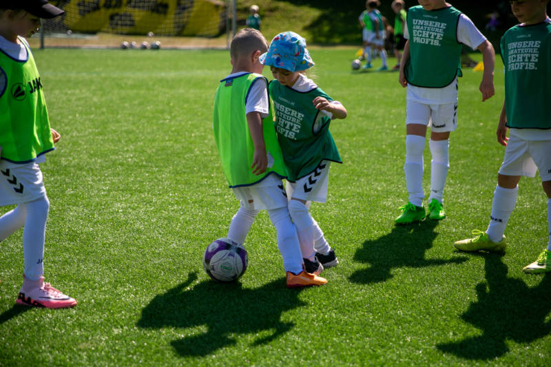 Zwei kleine Kinder kämpfen bei sonnigem Wetter spielerisch um den Ball auf einem grünen Kunstrasenplatz. Beide tragen Sportkleidung und Leibchen, eines mit einem bunten Sonnenhut. Auf den grünen Leibchen steht der Schriftzug „Unsere Amateure. Echte Profis.“. Weitere Kinder beobachten die Szene im Hintergrund, die eine fröhliche und aktive Atmosphäre im Kinderfußballtraining widerspiegelt.