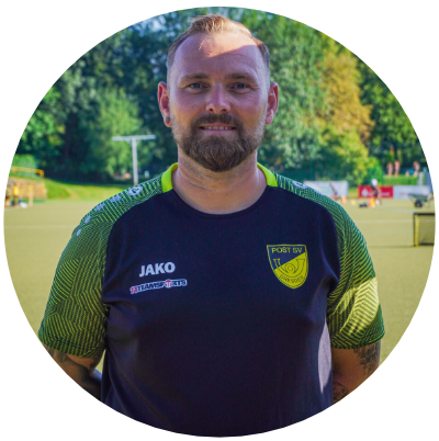 Ein Mann steht auf einem Fußballplatz und blickt freundlich in die Kamera. Er trägt ein dunkles Sportshirt mit gelben Akzenten und dem Logo von „Post SV Dresden“. Auf dem Shirt sind außerdem die Marken „JAKO“ und „Teamsport“ zu sehen. Im Hintergrund erkennt man Tore, Hütchen und grüne Bäume – typische Umgebung eines Fußballtrainings.