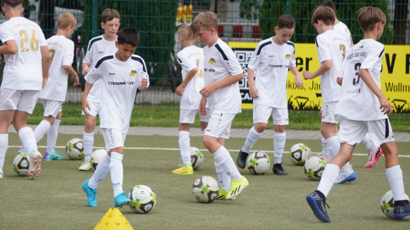 Mehrere Kinder in weißen Fußballtrikots mit dem Aufdruck „Post SV Dresden“ und „TeamSports“ trainieren auf einem Kunstrasenplatz. Jedes Kind hat einen eigenen Ball und führt Dribbelübungen aus. Im Vordergrund steht ein gelbes Markierungshütchen, im Hintergrund ein Zaun mit Werbebannern. Die Szene zeigt konzentriertes Training in einem Sommer-Fußballcamp.